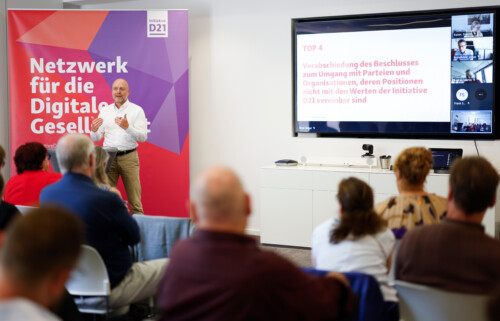 Eine Person redet an einem Pult zu anderen. Im hintergrund eine Präsentation und ein Banner der Initiative D21