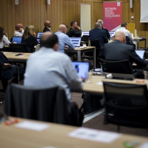 Aufnahme eines Tagungsraumes von hinten. In mehreren Tischreihen sitzen Menschen vor Laptops und schauen nach vorne, wo mit einem Beamer etwas an die Leinwand geworfen wird.