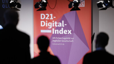 Ein Banner mit dem Schriftzug D21-Digital-Index. Jährliches Lagebild zur Digitalen Gesellschaft steht auf einer Bühne und wird angestrahlt. Im Vordergrund sieht man verschwommen Personen, die in Richtung des Banners gucken.